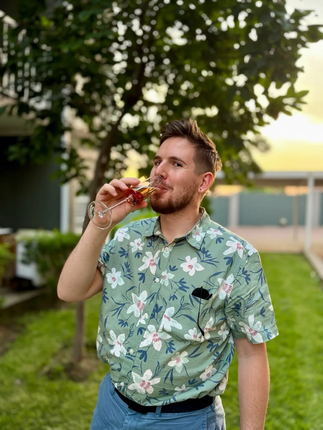 Cameron in a floral shirt sipping champagne in a garden at sunset
